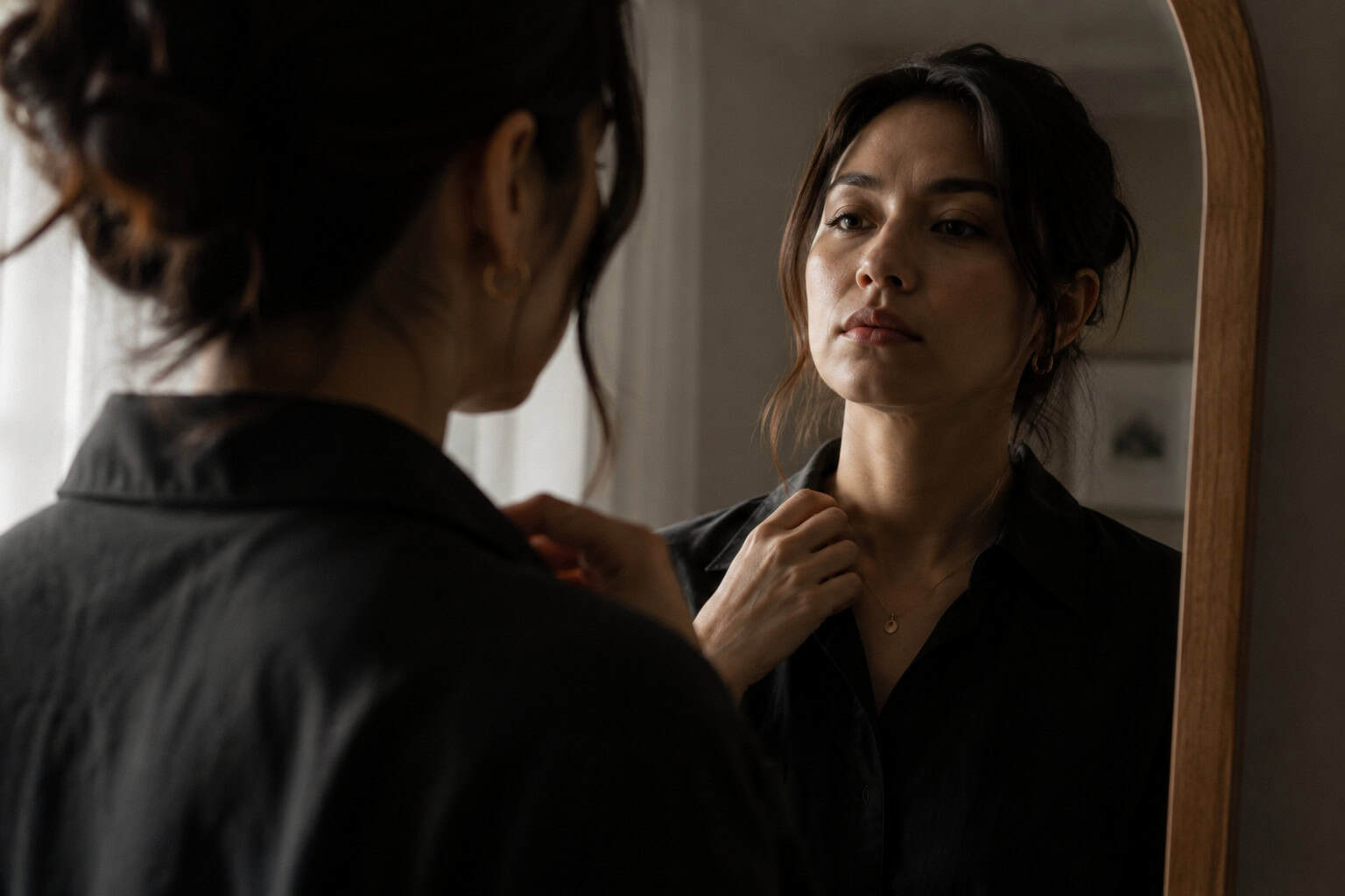 Person adjusting their posture in front of a mirror with face partially obscured by shadow, suggesting a contrast between outward composure and internal experience