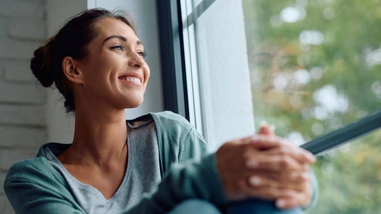 Smiling woman day dreaming while relaxing by the window at home, concept of contentment, happiness