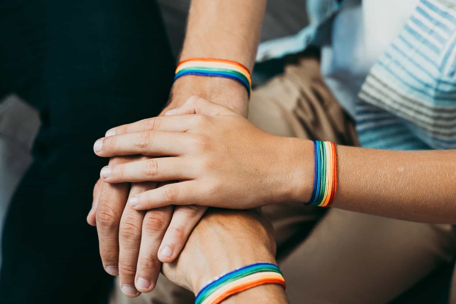 Three pairs of hands are stacked on top of each other, each wearing rainbow striped bracelets