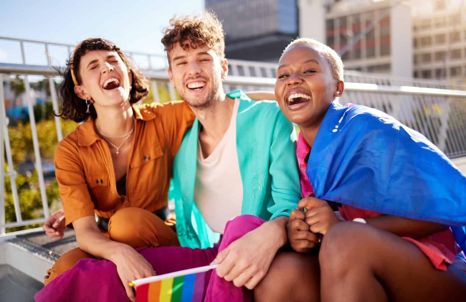 Three young adults are smiling and laughing on a sunny day, holding a pride flag. Concept of friendship, support, pride, sense of community