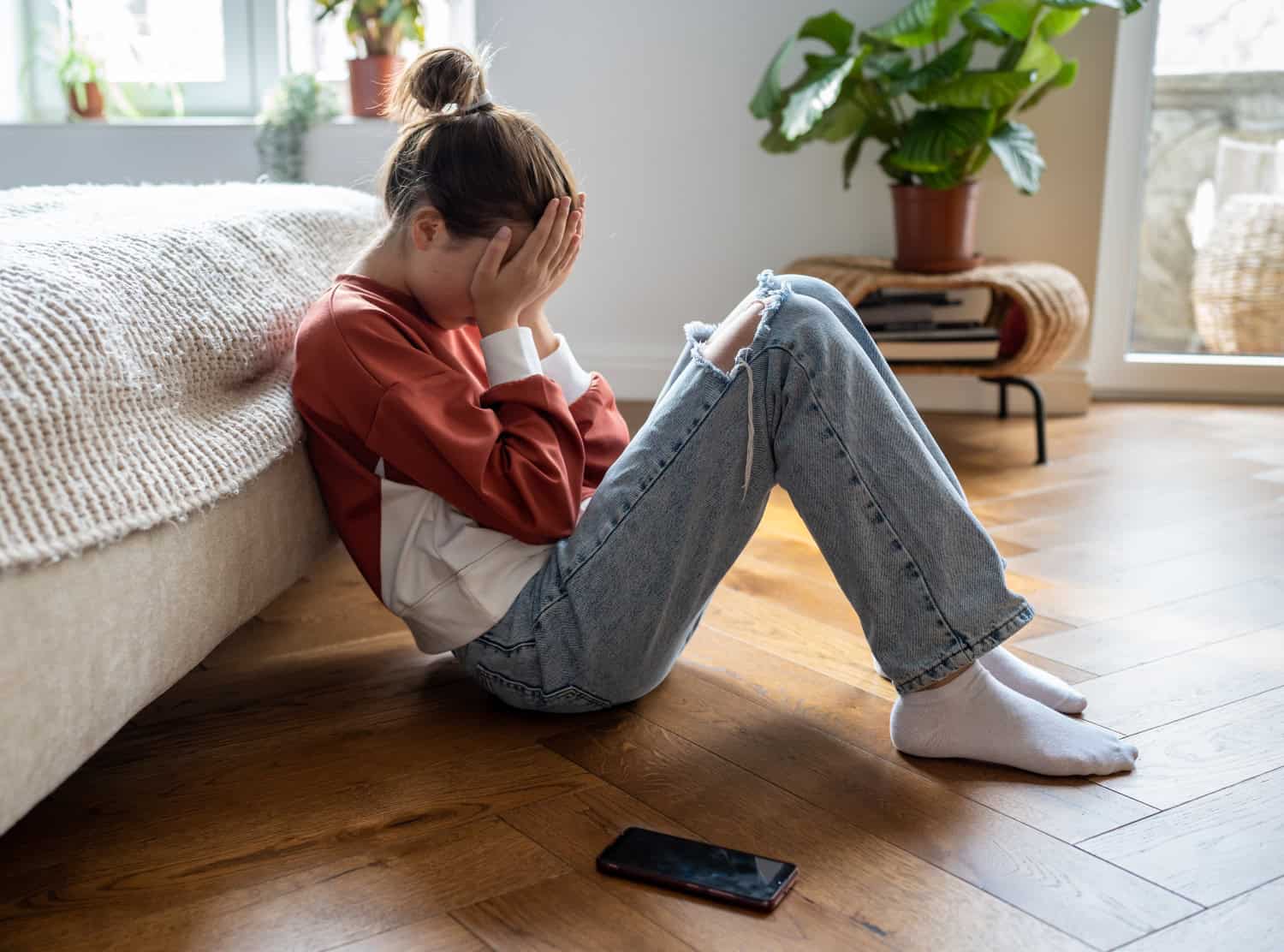 Unhappy teen girl covering face with hands crying while sitting on floor with mobile phone nearby