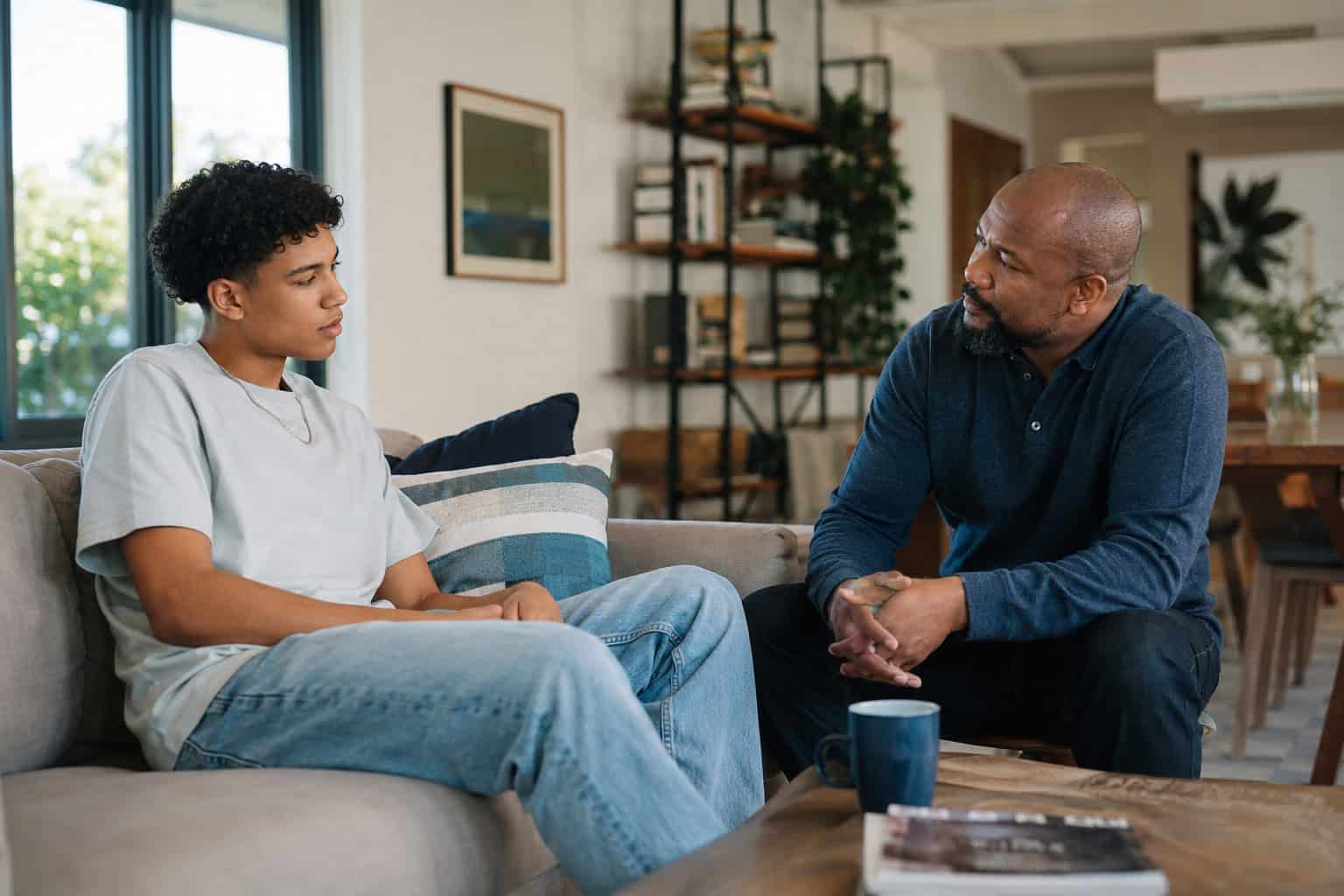a parent and young adult having a quiet, honest conversation together in a bright contemporary living room